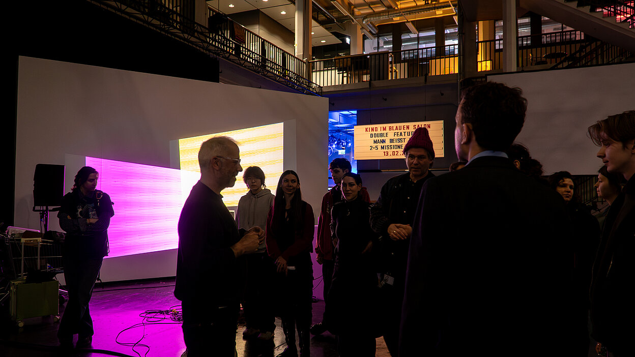 A group of people gathered around a man in glasses speaking at the Shifting Signals workshop in the HfG atrium with projected visuals behind them