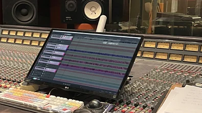 A computer monitor displaying a multi-track audio editing session sits on a large analog mixing console in a recording studio