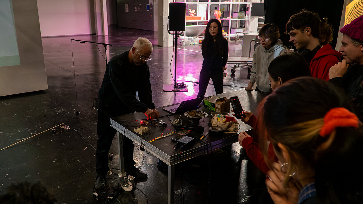A group of people gather around a table with electronic equipment and circuits while one person demonstrates during the Shifting Signals workshop